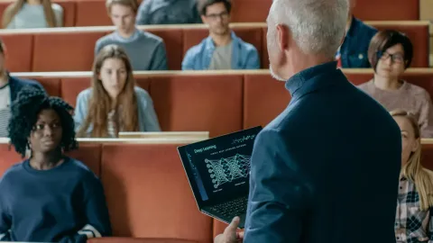 A professor is standing in front of a lecture hall, facing a group of students.